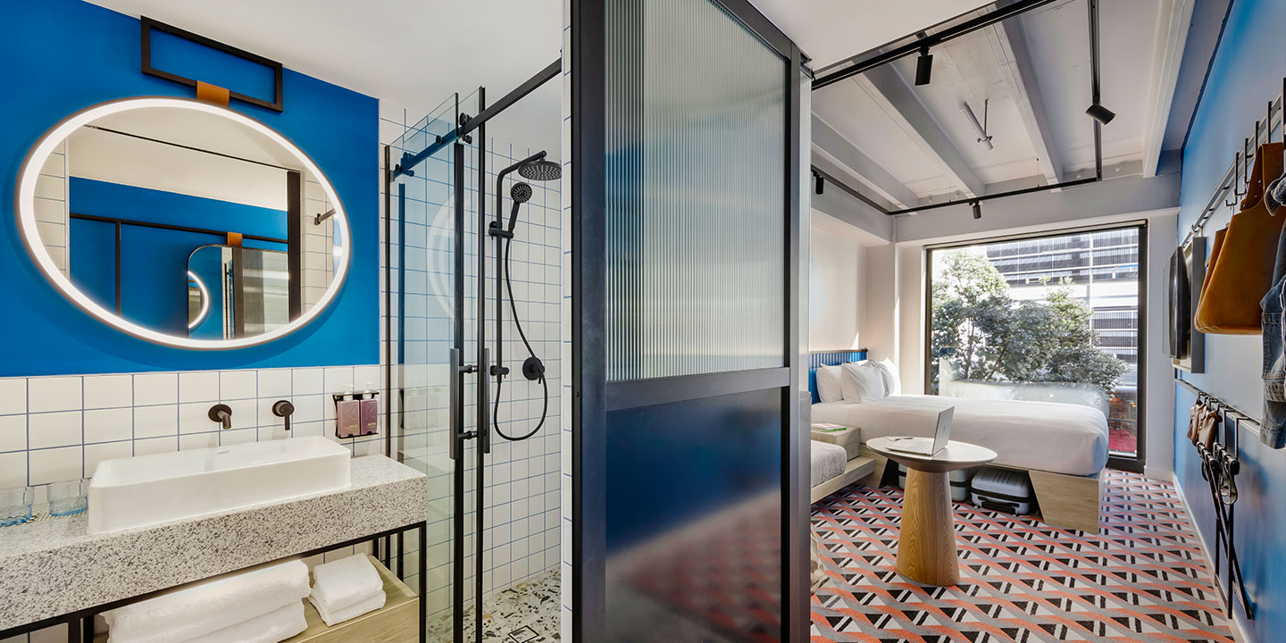 Modern hotel room with geometric decor: features a glass shower with black fixtures, a round mirror, a bed by a large window, and a patterned rug. Bright and inviting.