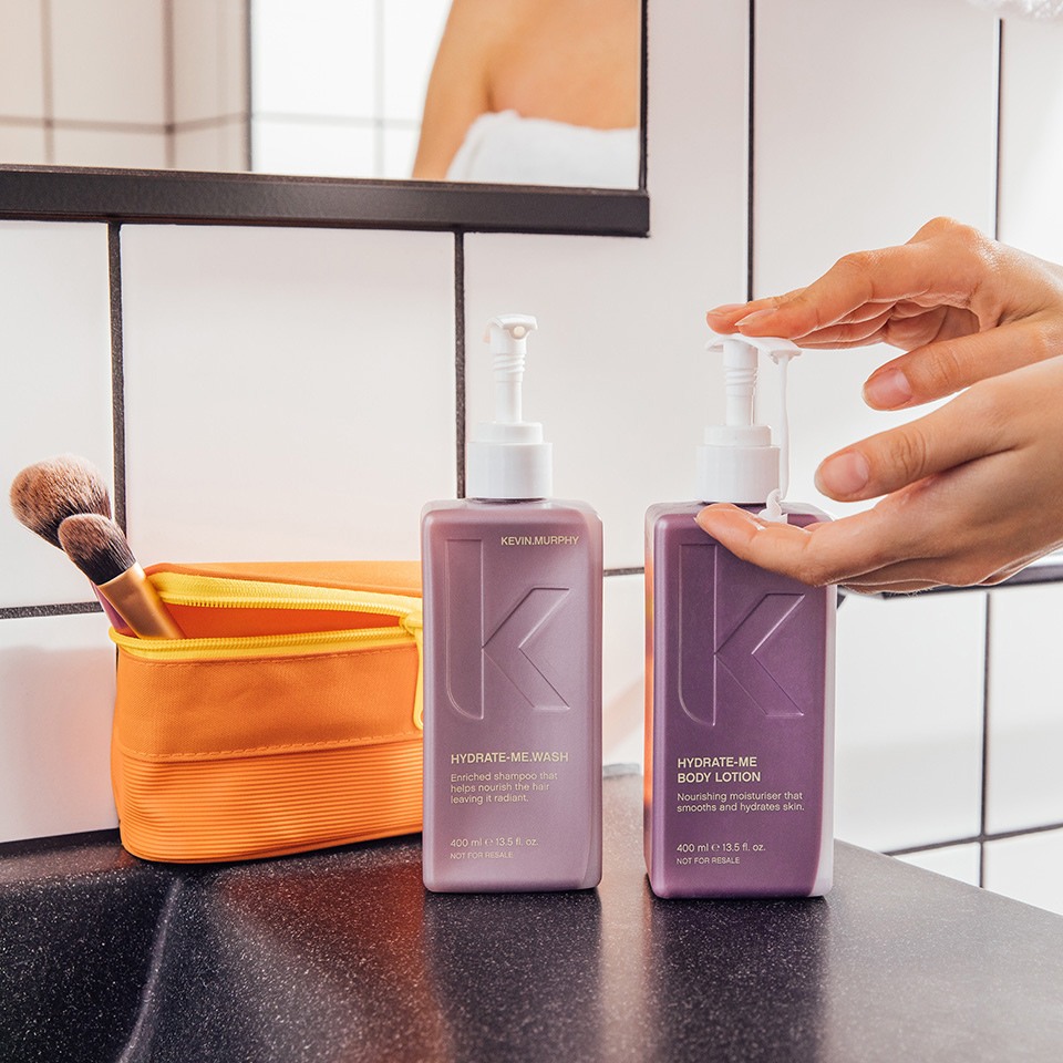 A hand pumps Kevin Murphy Hydrate-Me products; shampoo and body lotion on a bathroom counter, with a makeup brush and orange bag nearby.