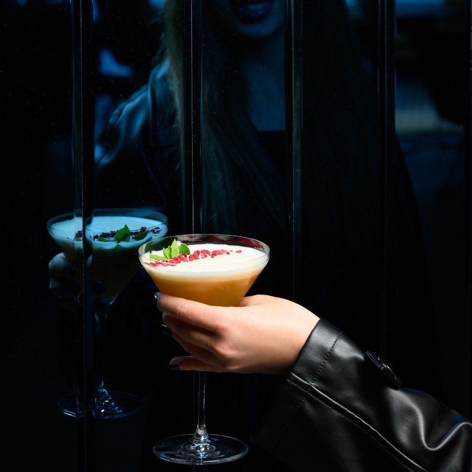 A hand holding a cocktail glass garnished with mint and red berries, reflected in a dark, shiny surface. Dim lighting and shadowy ambiance.